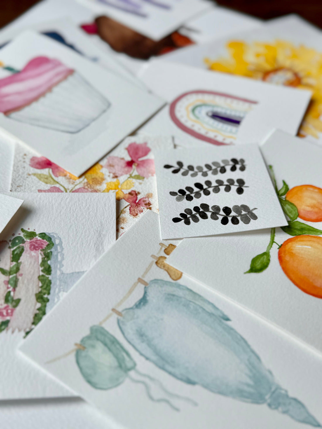 A colorful scatter of watercolor paintings including a baby gown, oranges, a rainbow and a yellow flower.