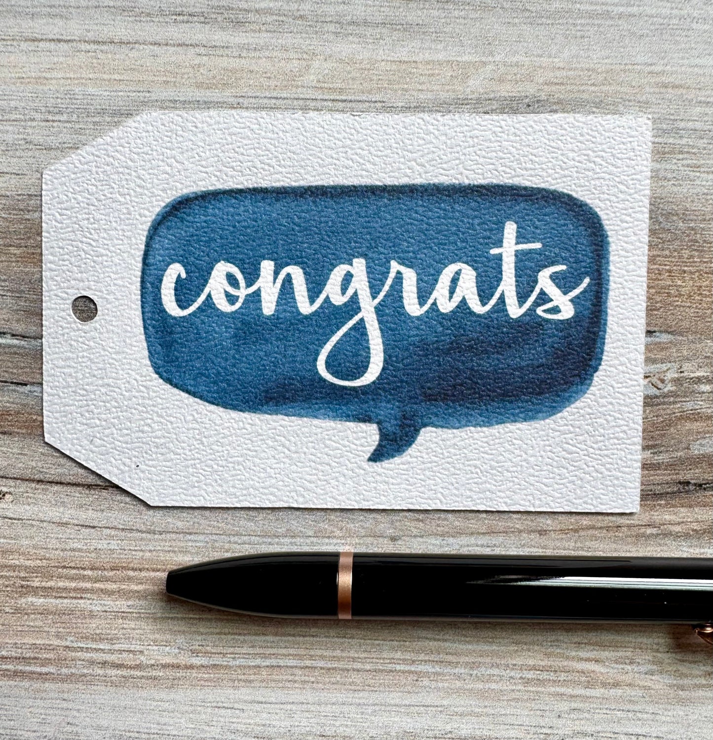 Gift Tag with blue speech bubble and 'congrats' text placed on a wooden surface with a pen.