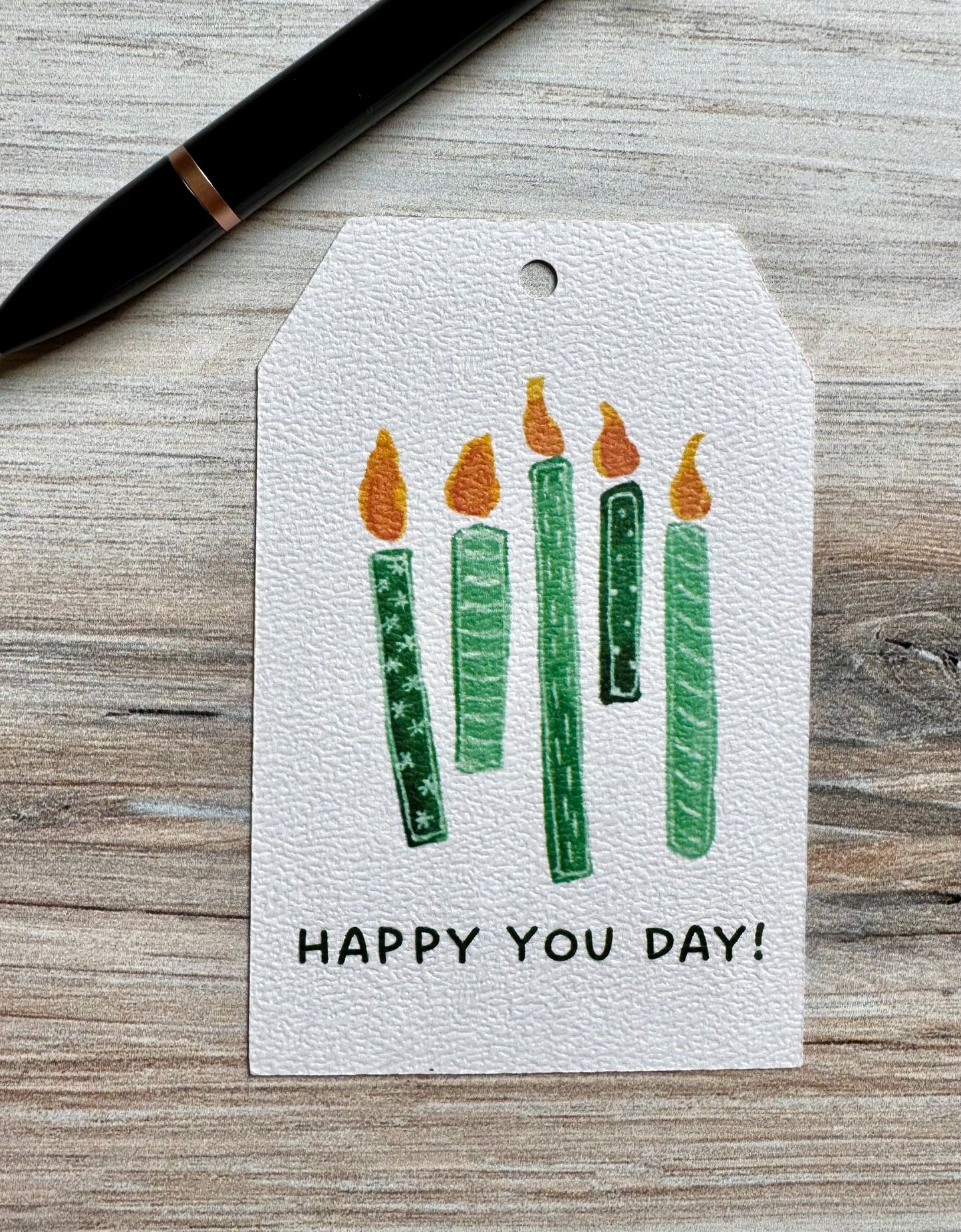 Gift tag with 5 green lit candles and 'Happy You Day!' text placed on a wooden surface