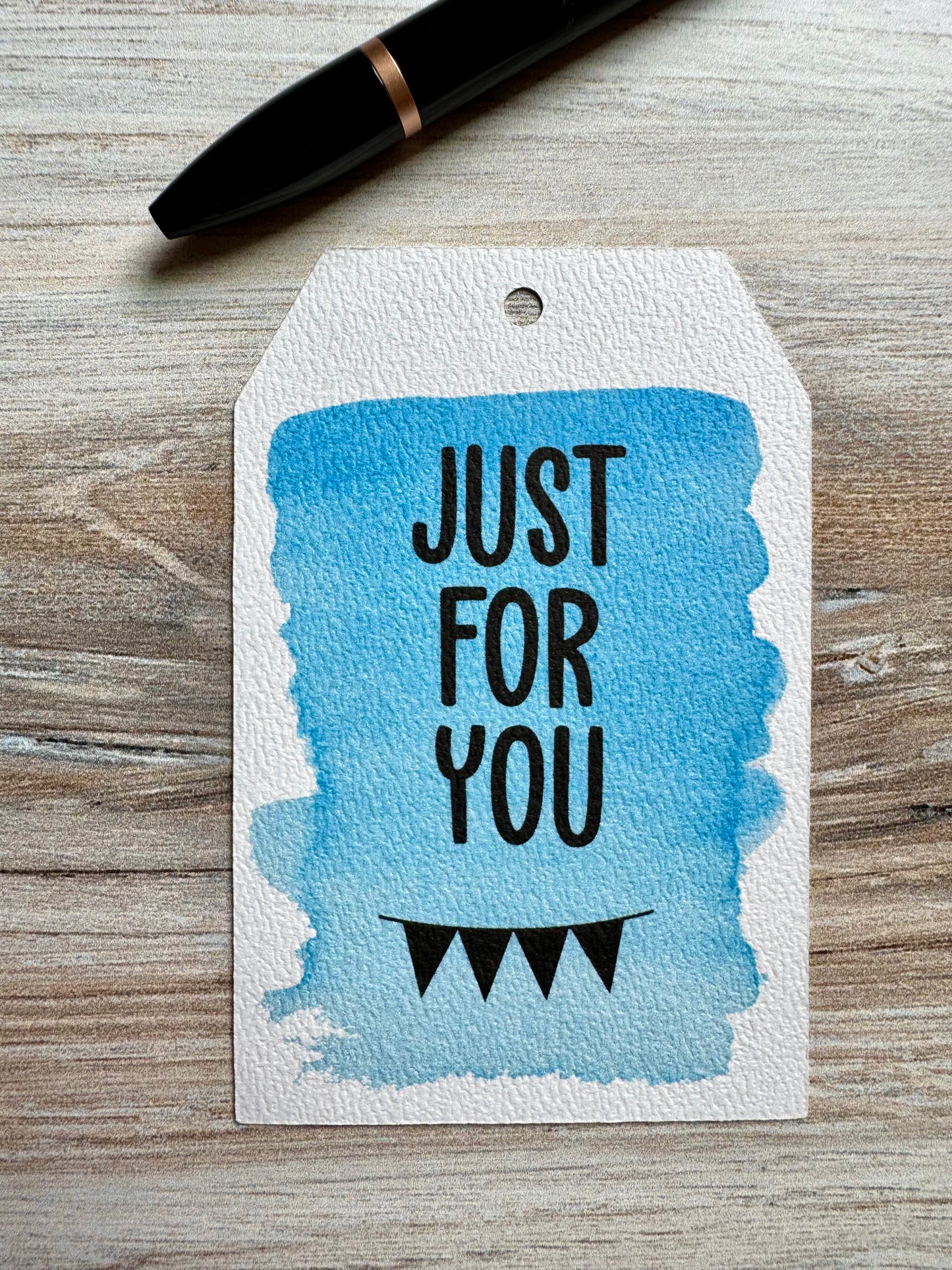 Gift tag with 'Just For You' printed over a watercolored blue background, placed on a wooden surface