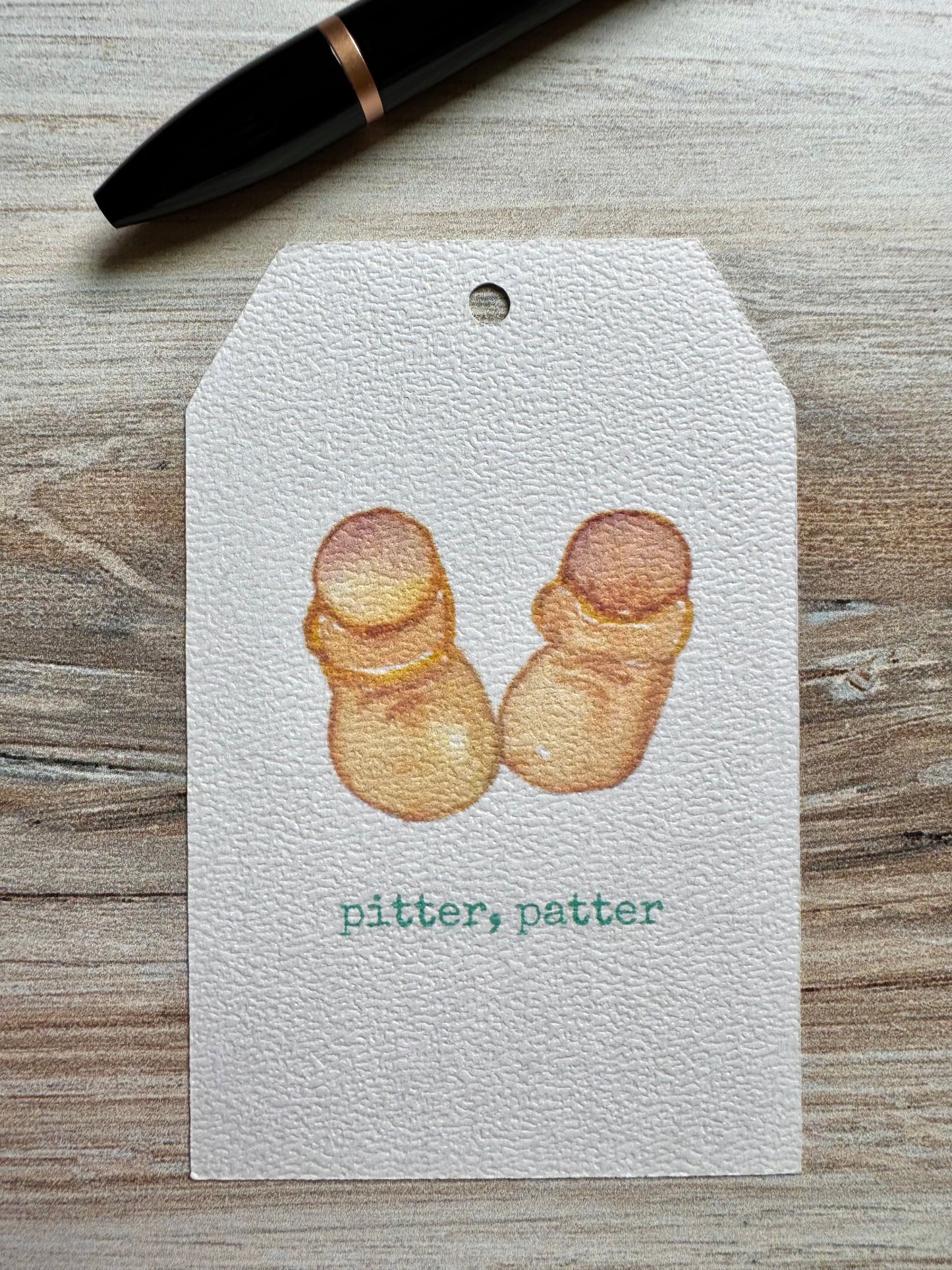 Gift Tag with baby booties illustration and 'pitter, patter' text placed on a wooden surface with a pen.