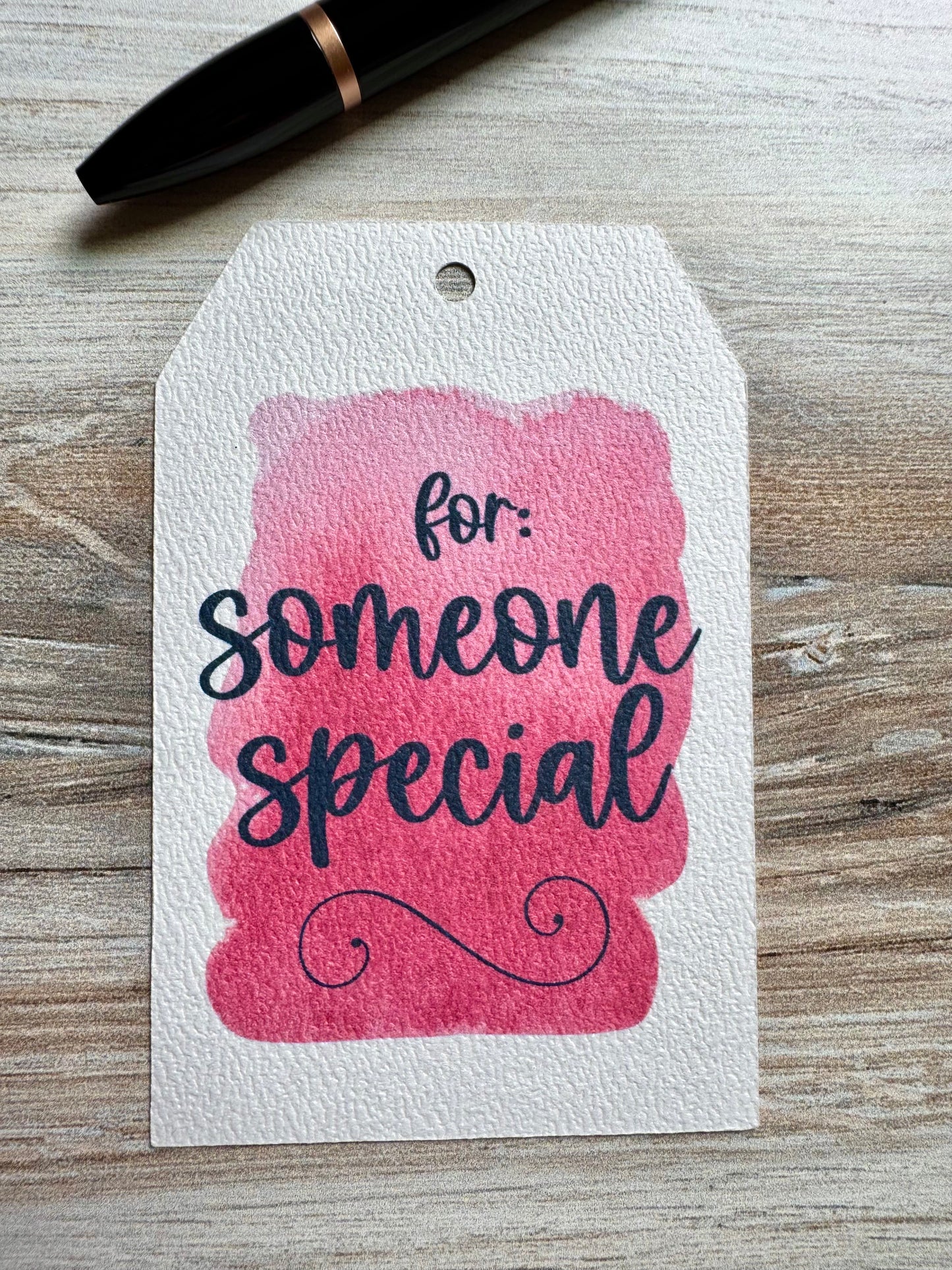 Gift Tag with pink watercolor background, and the words "for someone special" printed over it. Placed on a wooden space with a pen above it.