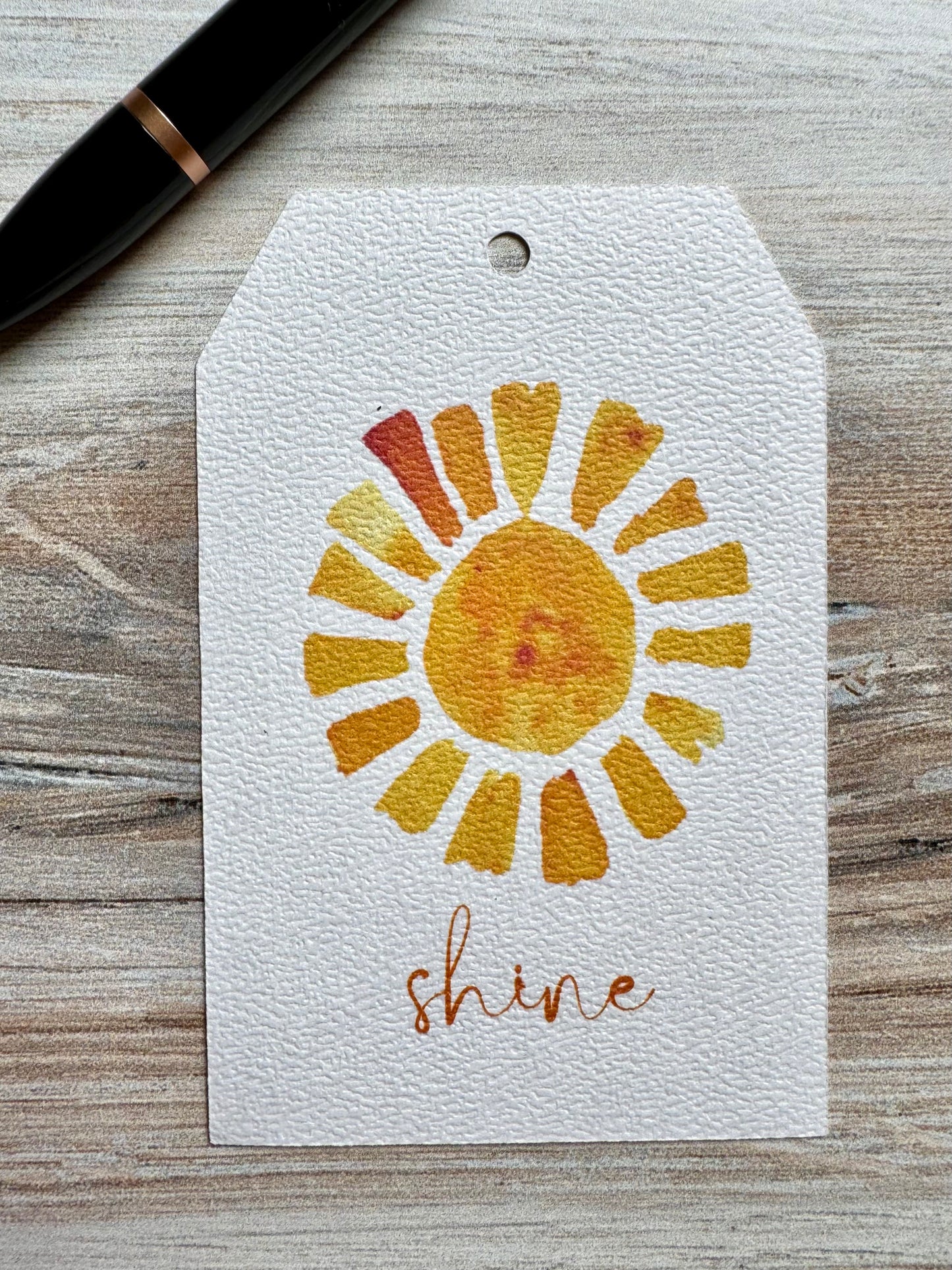 Gift tag with a watercolor sun design and the word 'shine' placed on a wooden surface