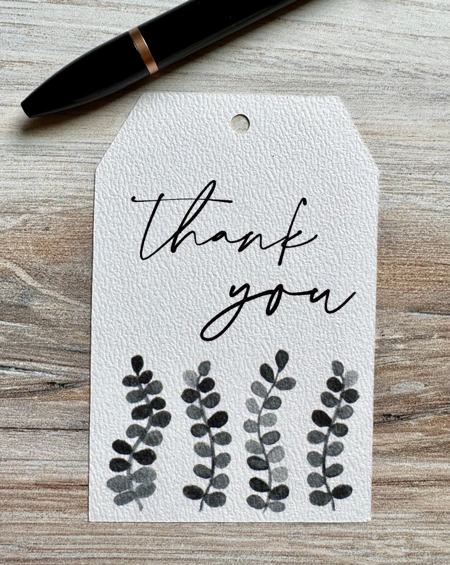 White gift tag with 'Thank you' text and grayscale leaf designs placed on a wooden surface