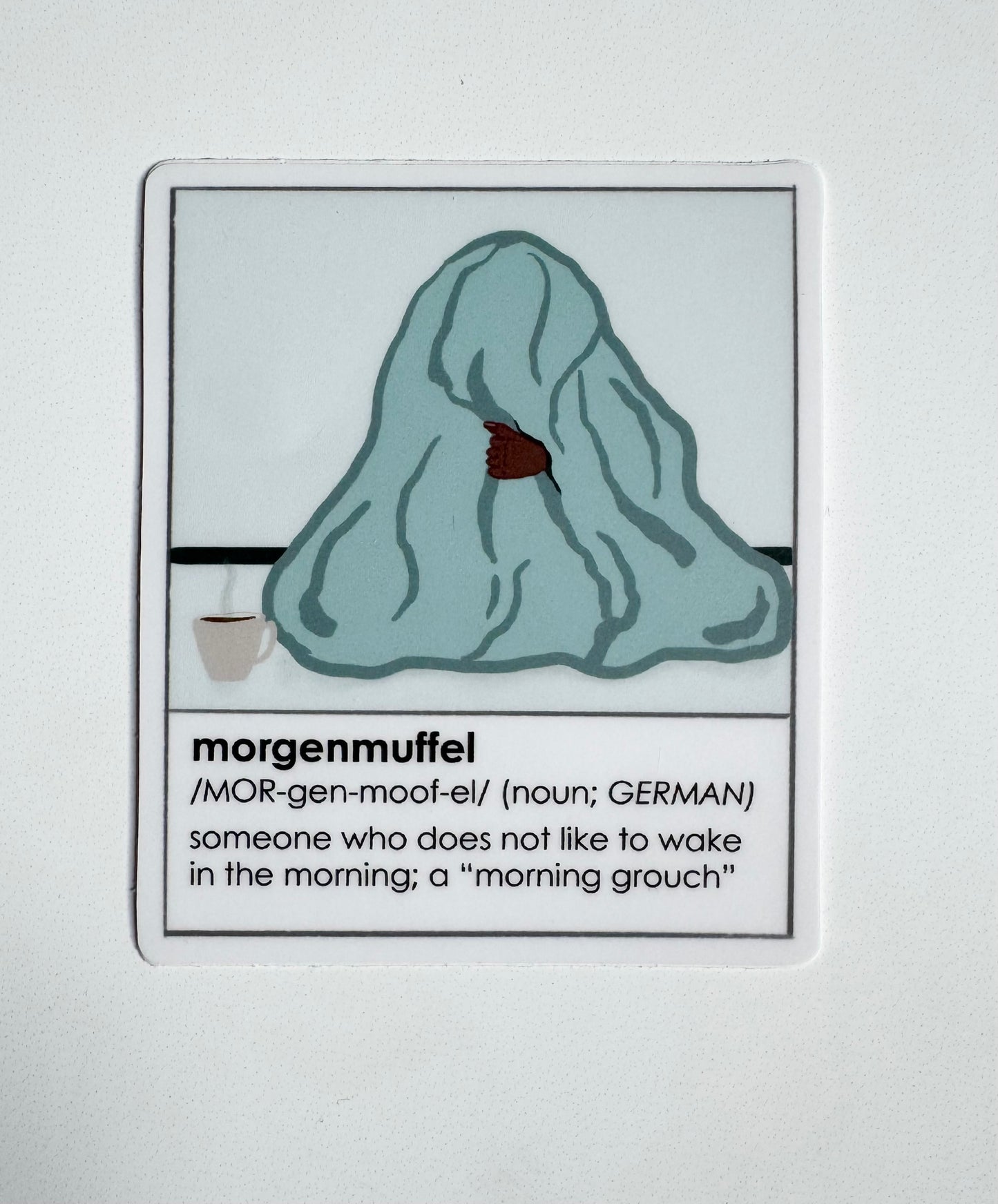 Sticker with illustration of a person with dark skin under a blanket and definition of 'morgenmuffel' on a white background