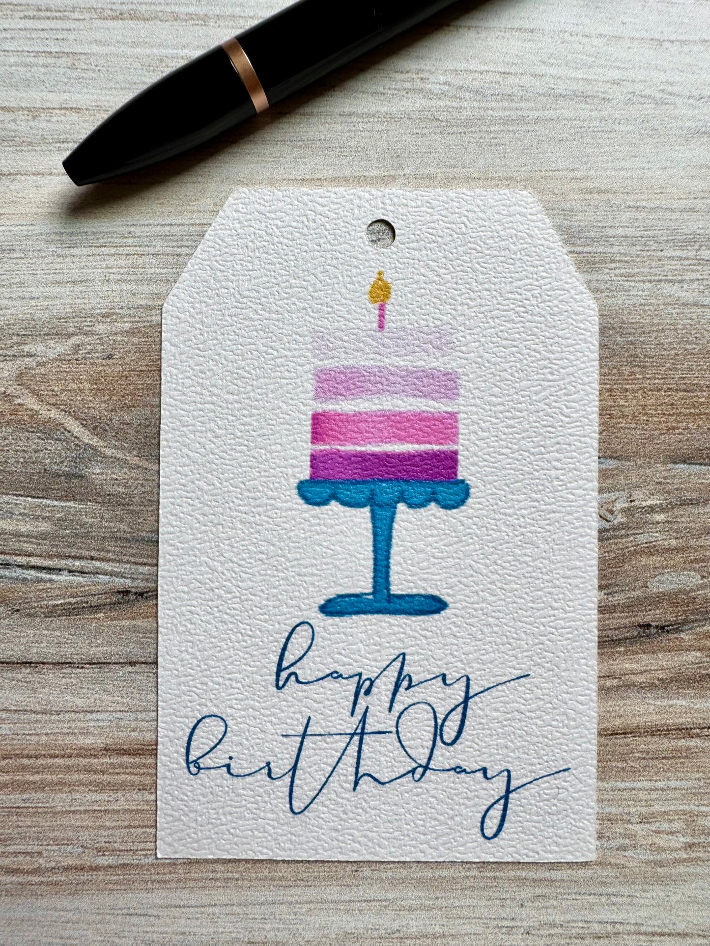 Gift tag with birthday cake  and a lit candle illustration and 'happy birthday' script, placed on a wooden surface.