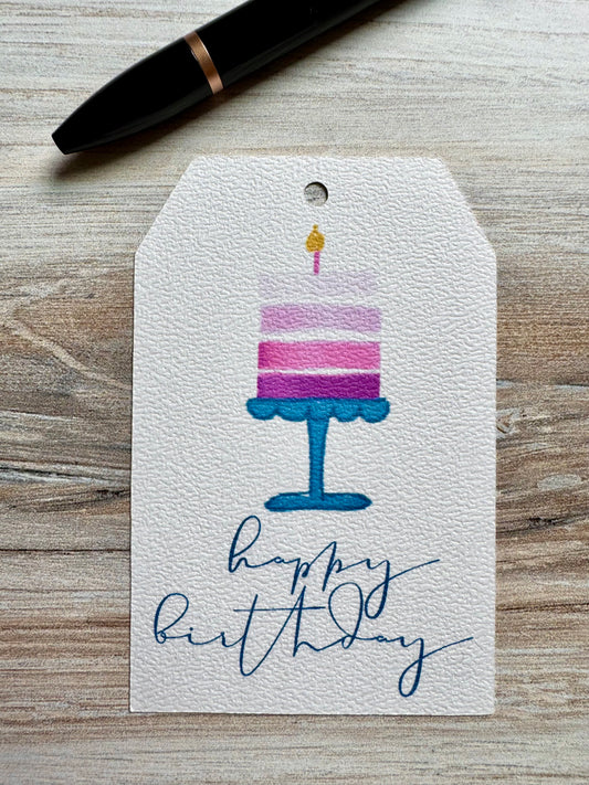 Gift tag with birthday cake  and a lit candle illustration and 'happy birthday' script, placed on a wooden surface.