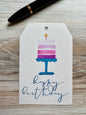 Gift tag with birthday cake  and a lit candle illustration and 'happy birthday' script, placed on a wooden surface.