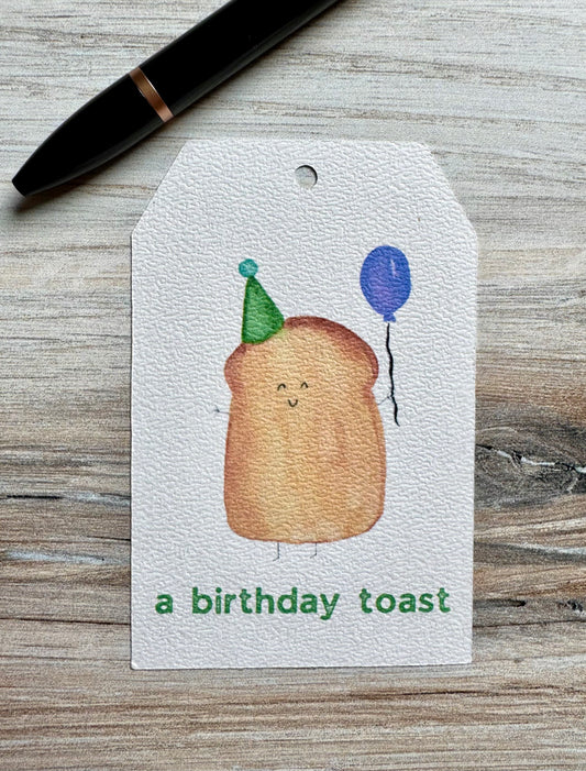 Gift tag with a cartoon toast wearing a party hat and balloon placed on a wooden surface.