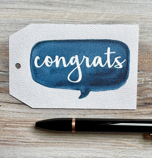 Gift Tag with blue speech bubble and 'congrats' text placed on a wooden surface with a pen.