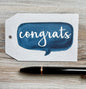 Gift Tag with blue speech bubble and 'congrats' text placed on a wooden surface with a pen.