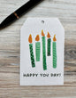 Gift tag with 5 green lit candles and 'Happy You Day!' text placed on a wooden surface