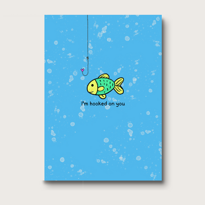 Digital illustration of a fish swimming just below a fishing hook with a heart-shaped tip. Below the fish, the text reads "I'm hooked on you." A charming and romantic design perfect for love or Valentine's Day cards. 