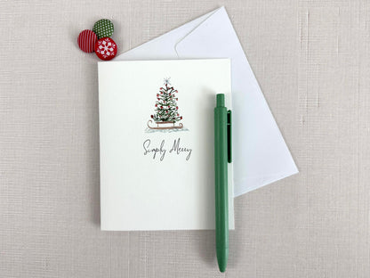 Christmas card with a tree design that reads "Simply Merry", pictured with a  pen, and envelope on a textured surface