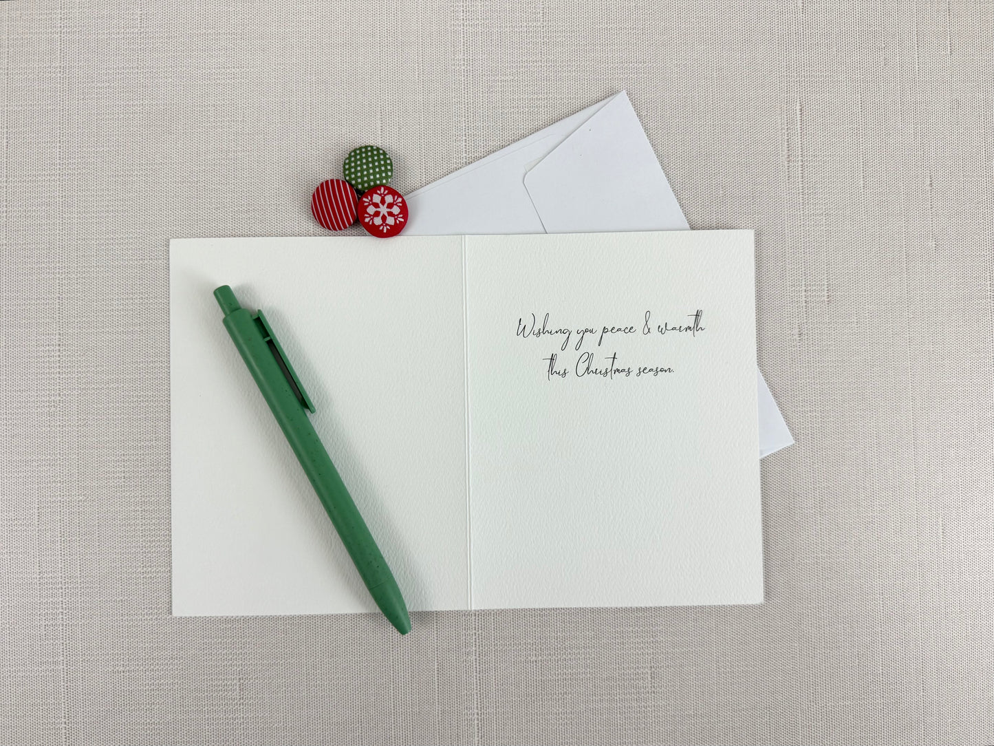 Open card with Christmas message that reads "Wishing you peace & warmth this Christmas season.", a  pen, and decorative pins on a textured surface