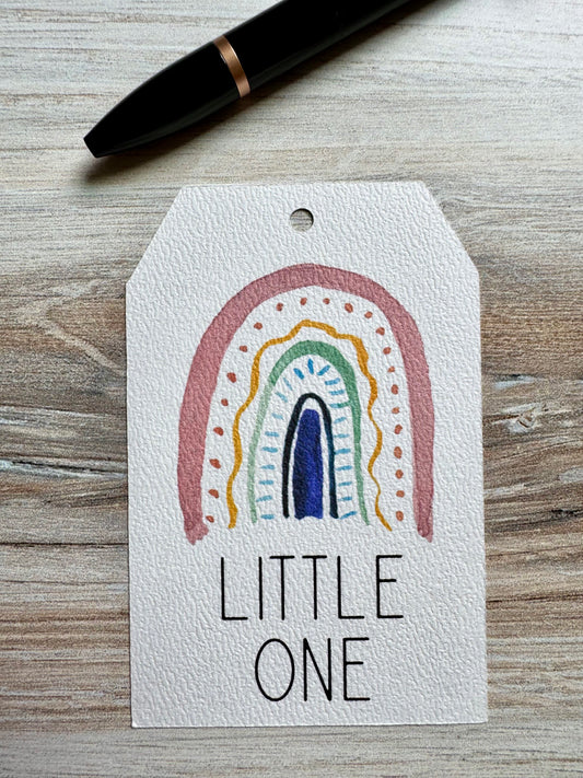 Gift tag with minimalist rainbow design and 'LITTLE ONE' text placed on a wooden surface