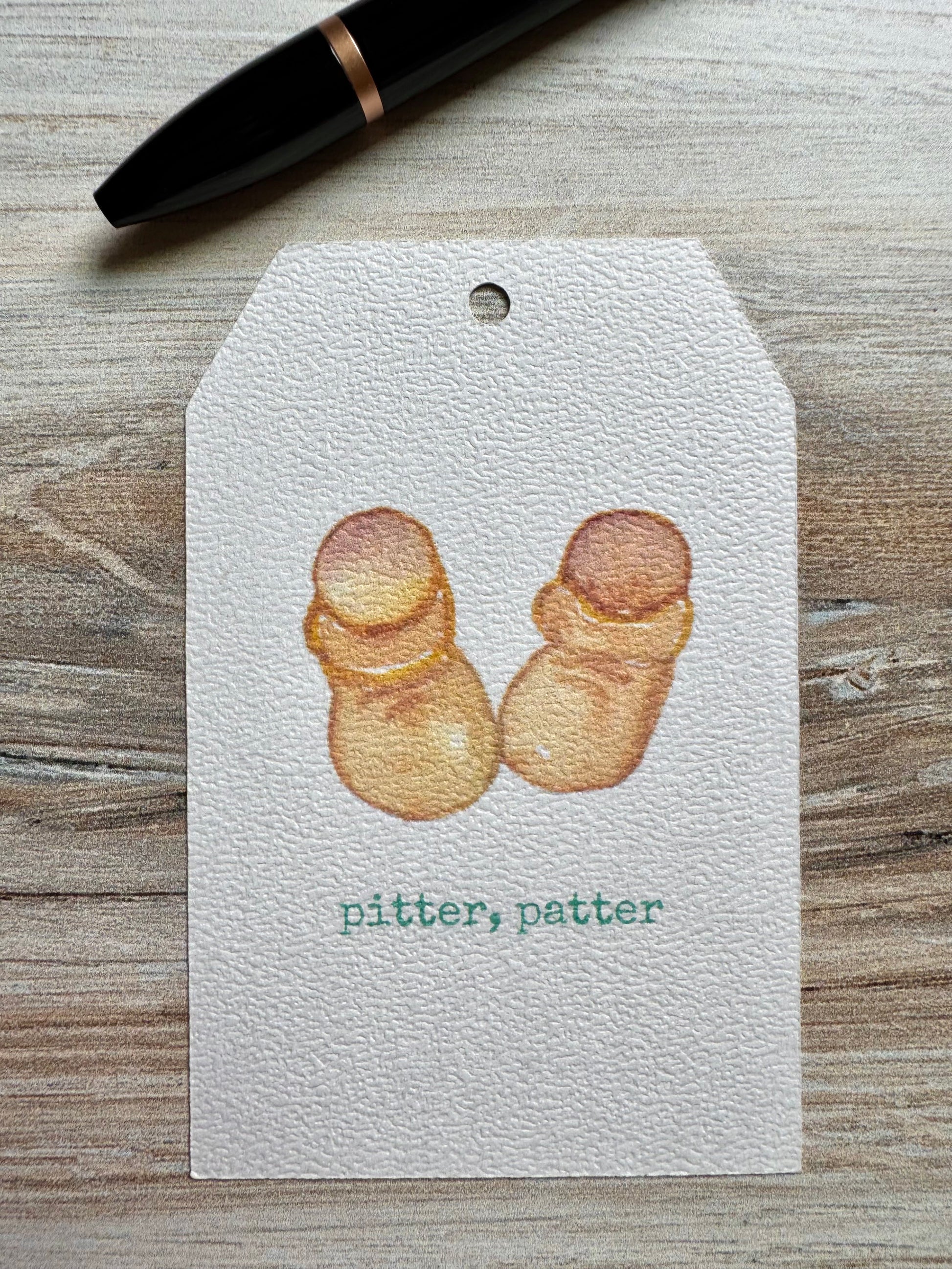 Gift Tag with baby booties illustration and 'pitter, patter' text placed on a wooden surface with a pen.