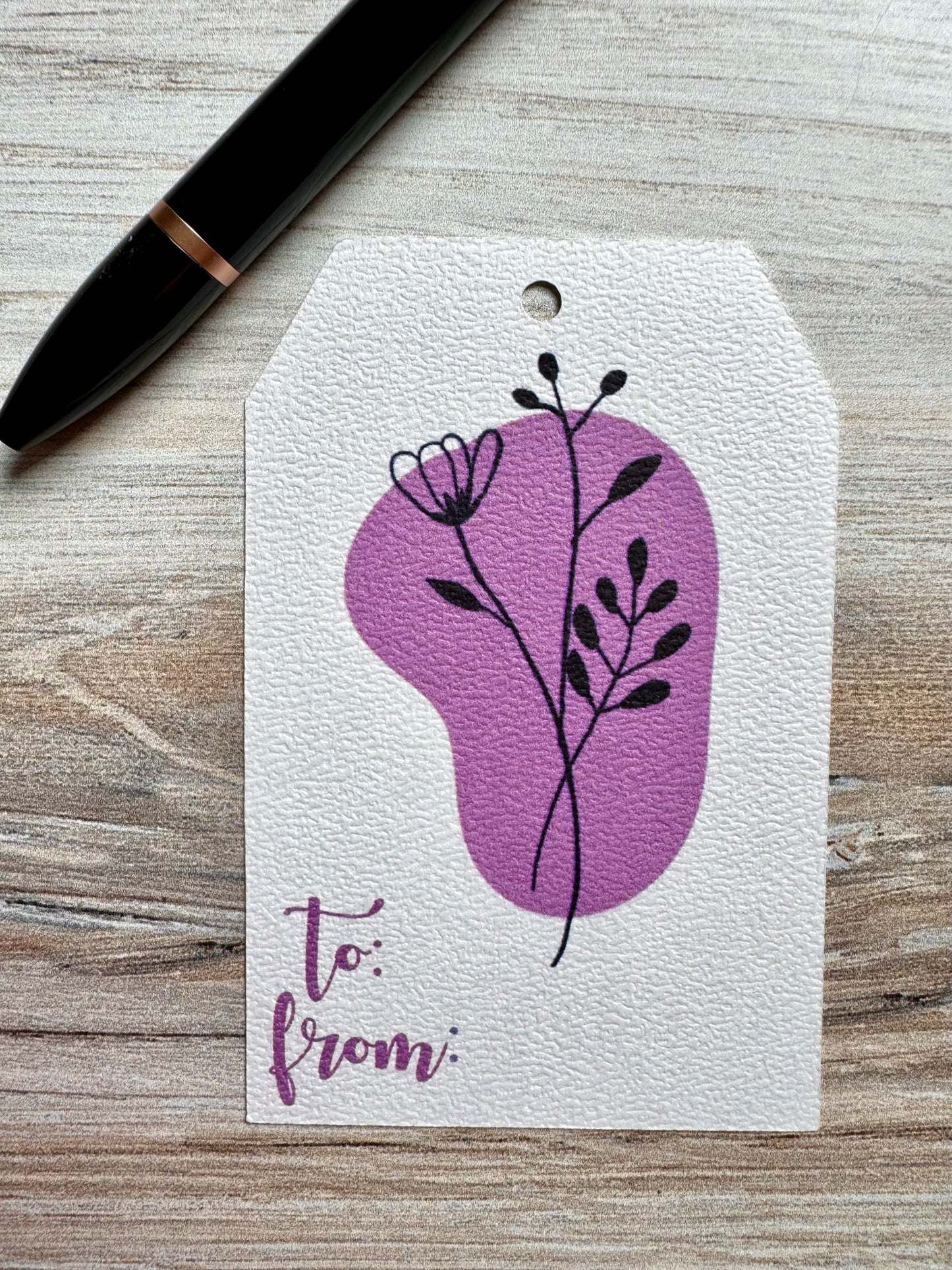 Gift tag with purple floral design and 'to/from' text on a wooden surface with a pen.