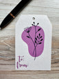 Gift tag with purple floral design and 'to/from' text on a wooden surface with a pen.