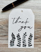White gift tag with 'Thank you' text and grayscale leaf designs placed on a wooden surface